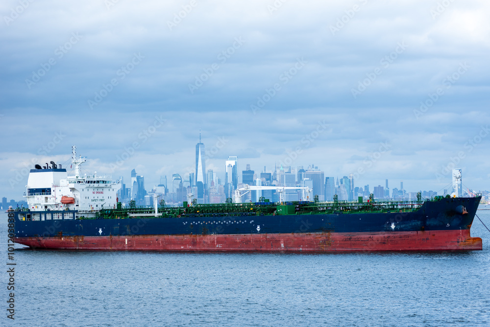 Fototapeta premium A cargo ship rests anchored in the bay, dominating the foreground. In the distance, the steel-blue skyline of New York City looms under a cloudy sky, blending into the moody, overcast atmosphere.