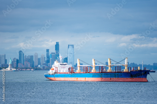 A cargo ship rests anchored in the bay, dominating the foreground. In the distance, the steel-blue skyline of New York City looms under a cloudy sky, blending into the moody, overcast atmosphere.