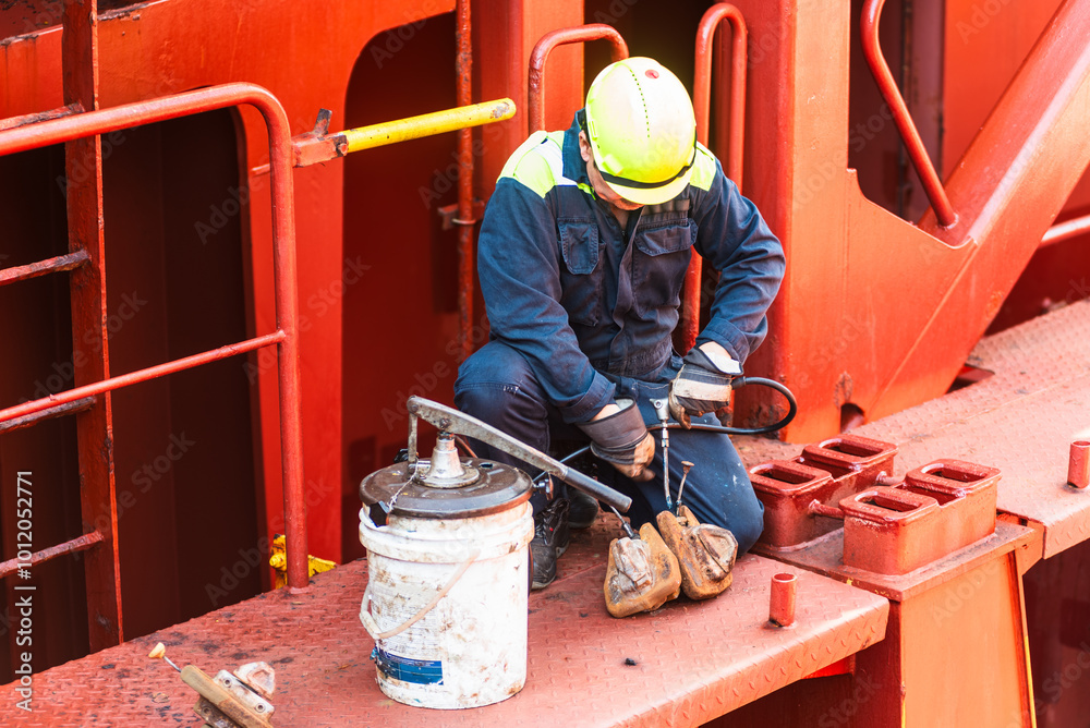 A crew member is crouched on the deck of a merchant ship, applying grease to container securing equipment. Focused on ensuring the safe and smooth operation of the gear during the voyage. A crew member is crouched on the deck of a merchant ship, applying grease to container securing equipment. Focused on ensuring the safe and smooth operation of the gear during the voyage.