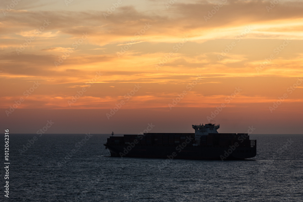 The calm silhouette of a massive container ship gliding across the smooth sea, enveloped by the golden hues of the sky as the sun bids farewell to the day. The calm silhouette of a massive container ship gliding across the smooth sea, enveloped by the golden hues of the sky as the sun bids farewell to the day.