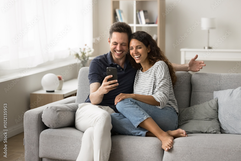 Cheerful married couple using application on mobile phone for Internet communication, sitting together on couch at home, having fun, smiling, laughing, watching online content on smartphone Cheerful married couple using application on mobile phone for Internet communication, sitting together on couch at home, having fun, smiling, laughing, watching online content on smartphone
