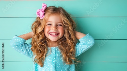 A cheerful little girl with curly hair and a pink bow, smiling exuberantly in front of a teal background, wearing a blue polka dot dress, embodying joy. photo