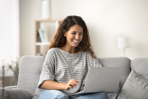 Happy beautiful Latin woman sitting on comfortable sofa with computer, typing on laptop, chatting, online, smiling, laughing, working on freelance business project