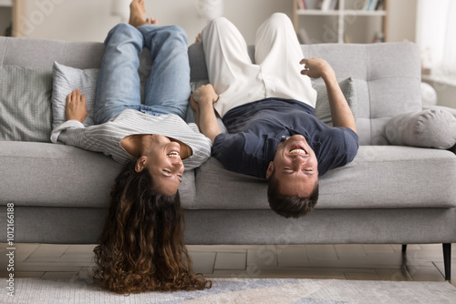 Cheerful cute young couple relaxing on grey soft couch with legs on back and hanging heads to floor, lying upside down, talking, laughing, smiling, having fun at home, enjoying funny leisure