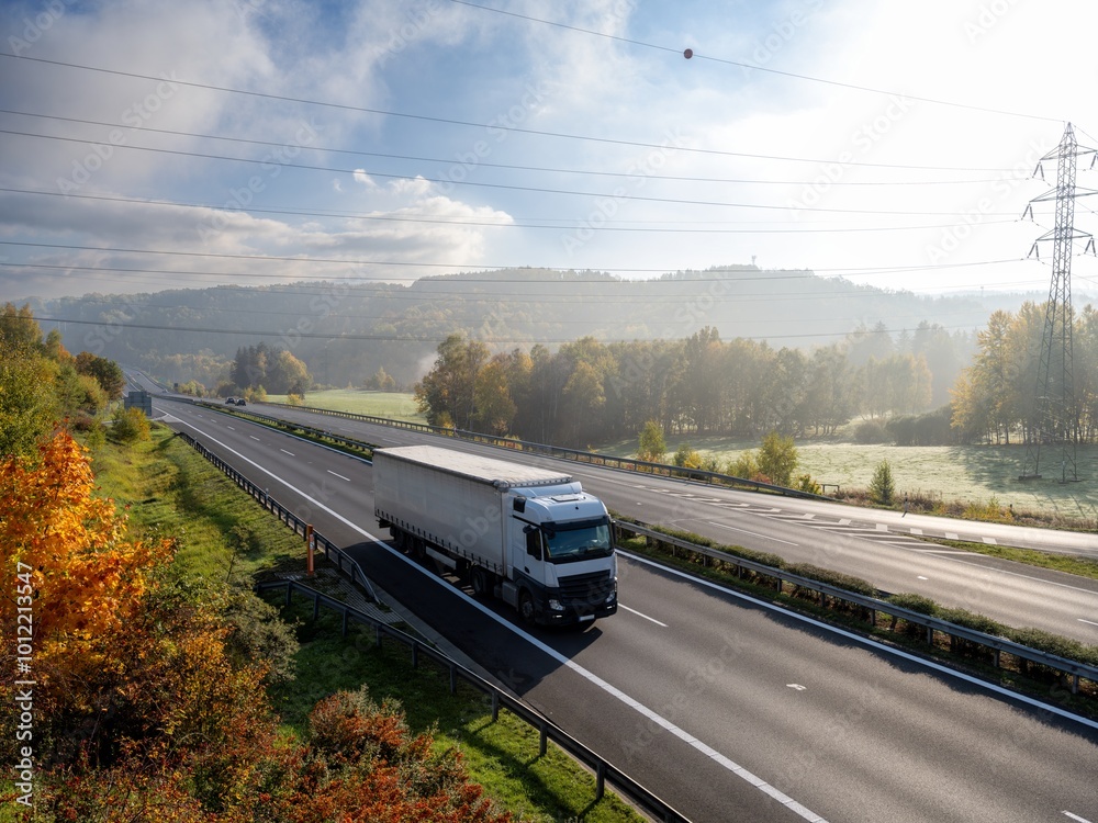 A white truck driving on an asphalt highway in a wooded landscape in autumn colors early in the morning
