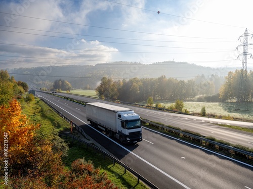 Obraz na płótnie A white truck driving on an asphalt highway in a wooded landscape in autumn colors early in the morning w salonie