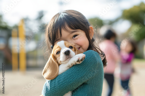 A cheerful preschooler with Japanese heritage, tightly hugging a fluffy beagle puppy on a playground. The joy on her face, with friends playing nearby, captures the essence of childhood happiness. photo