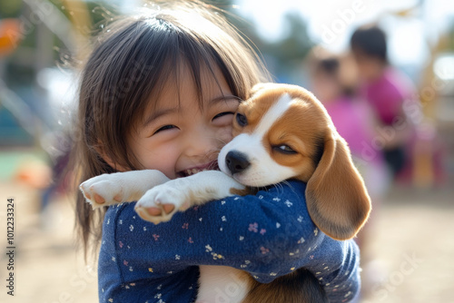 A cheerful preschooler with Japanese heritage, tightly hugging a fluffy beagle puppy on a playground. The joy on her face, with friends playing nearby, captures the essence of childhood happiness. photo