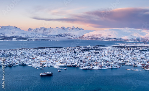 Aerial view of Tromso, Norway at sunset in winter from Storsteinen mountain. Tromsoya island with snowy roofs of Tromso downtown, harbor, cruise ship in fjord water and mountains in background