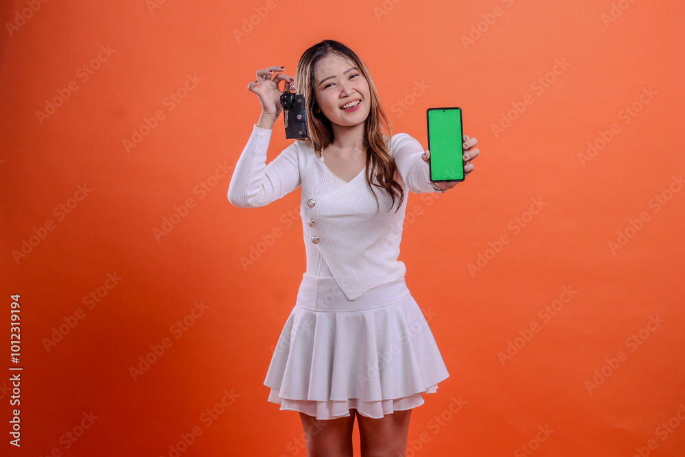 adult asian girl in white top standing cheerful hand holding car key and mobile phone screen, gadget in front isolated orange background. for transportation, rental services, vehicle business adult asian girl in white top standing cheerful hand holding car key and mobile phone screen, gadget in front isolated orange background. for transportation, rental services, vehicle business