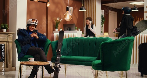 Asian tourist messaging on smartphone, asking friends to meet skiing on alpine slopes at mountain lodging complex. Winter sport fan checking phone and social media page before sliding down.