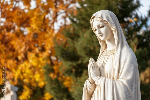 A serene statue of a woman with clasped hands in a colorful autumn setting.