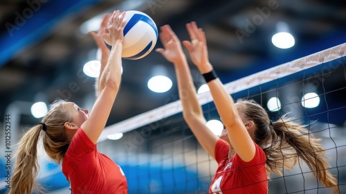Two female volleyball players jumping to intercept the ball at the net with intensity and focus, emphasizing competition and teamwork in an indoor sports setting. photo
