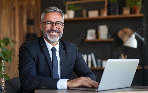 A smiling man in a suit sits at a desk with a laptop, surrounded by plants and books, exuding a professional and friendly atmosphere.