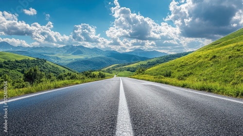 An asphalt road stretching through green mountain terrain, with a sunny sky filled with fluffy white clouds.