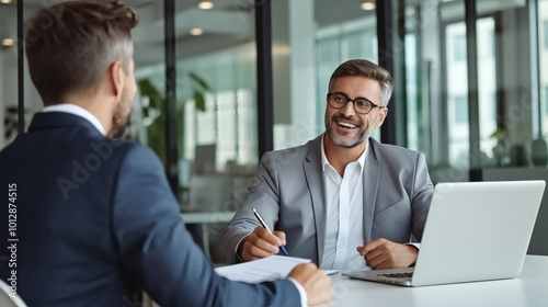 A professional and confident manager is having an interview with another man in the office, sitting at his desk while holding documents. 