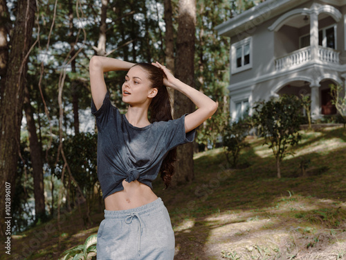 Relaxed young woman enjoying nature outdoors, wearing casual clothing, posing playfully with a background of lush greenery and a beautiful house nearby