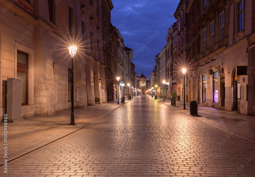 Naklejka premium Florianska Street in Krakow, Poland, photographed at dawn. The empty cobblestone street is lined with historic buildings, leading to the illuminated Florianska Gate in the distance