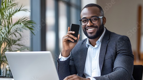 Smiling Black Man Holding a Smartphone While Working on a Laptop