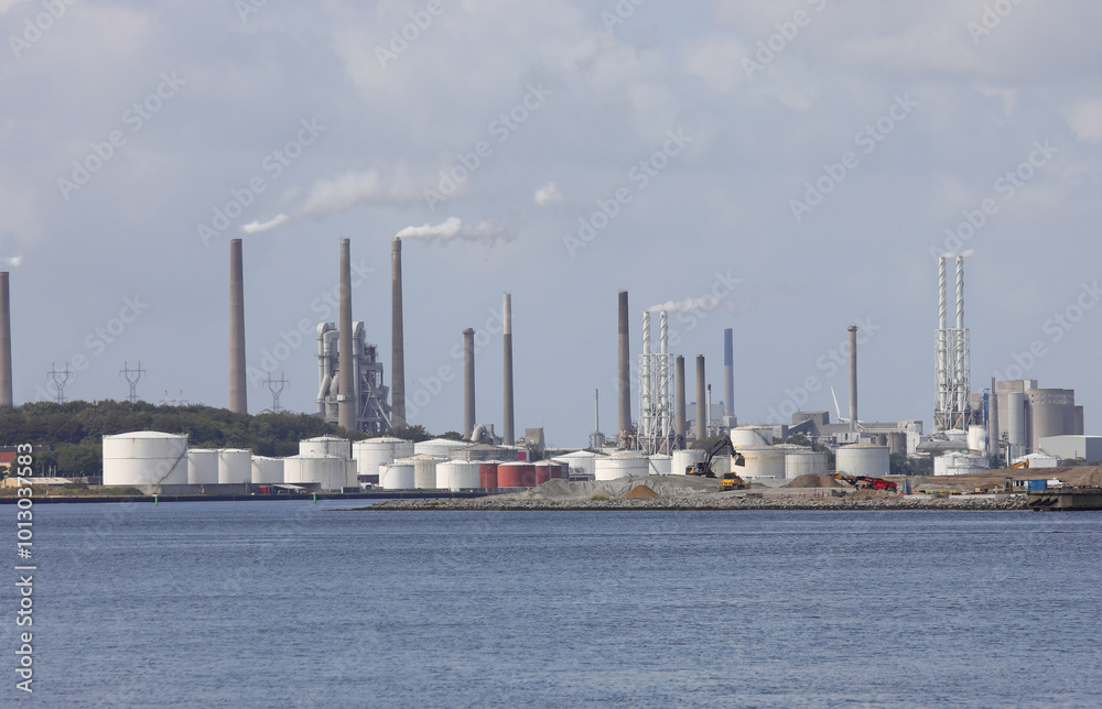 industrial plants and silos in a large industrial area near the sea with white polluting rising from tall chimneys industrial plants and silos in a large industrial area near the sea with white polluting rising from tall chimneys