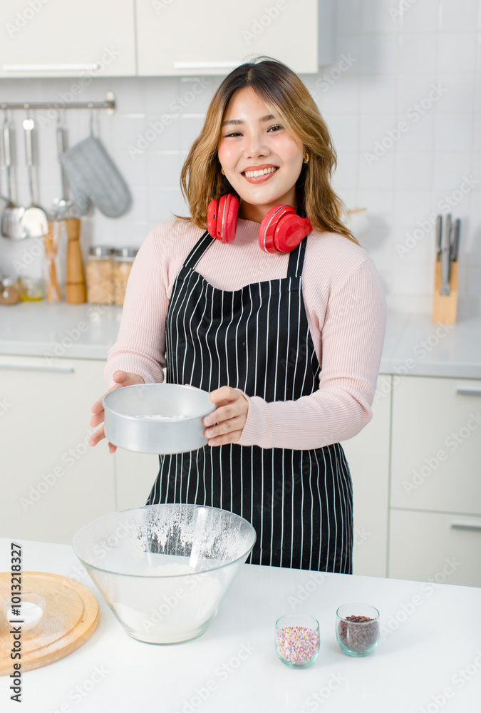Young happy Asian beautiful woman, female wearing aprons intends and headset standing in kitchen sifting flour for homemade bakery, bread baking making pastry or cake. Lifestyle enjoy cooking at home Young happy Asian beautiful woman, female wearing aprons intends and headset standing in kitchen sifting flour for homemade bakery, bread baking making pastry or cake. Lifestyle enjoy cooking at home