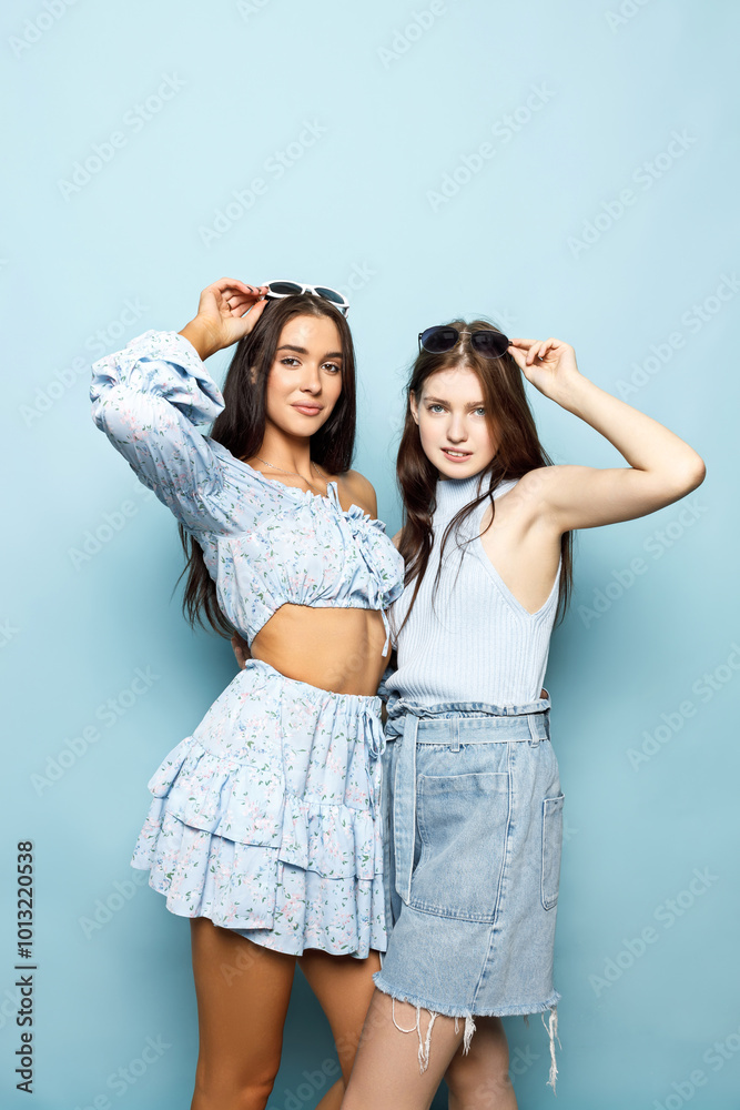 Two Friends Pose Cheerfully Against a Light Blue Backdrop, Showcasing Trendy Outfits and Sunglasses in a Lively Indoor Setting Two Friends Pose Cheerfully Against a Light Blue Backdrop, Showcasing Trendy Outfits and Sunglasses in a Lively Indoor Setting
