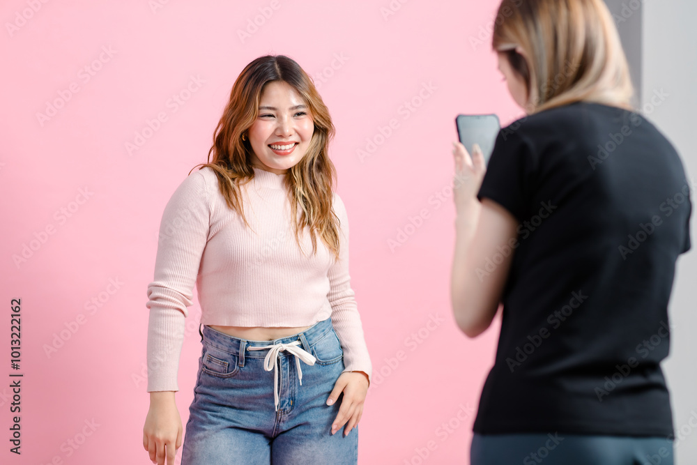 Two funny happy friends, Asian young woman pretty cheerful female smiling attractive enjoying holding a mobile phone and taking photo of friend poses isolated pastel over pink color wall background. Two funny happy friends, Asian young woman pretty cheerful female smiling attractive enjoying holding a mobile phone and taking photo of friend poses isolated pastel over pink color wall background.