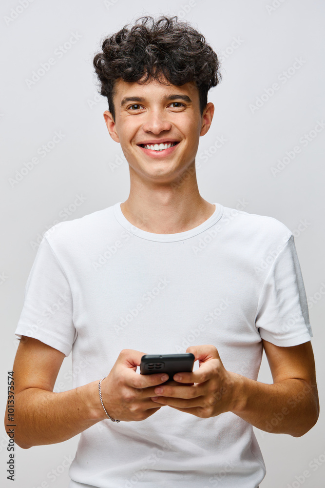 Young man smiling while holding a smartphone, dressed in a white shirt, against a soft gray background, conveying a sense of happiness and engagement with technology Young man smiling while holding a smartphone, dressed in a white shirt, against a soft gray background, conveying a sense of happiness and engagement with technology