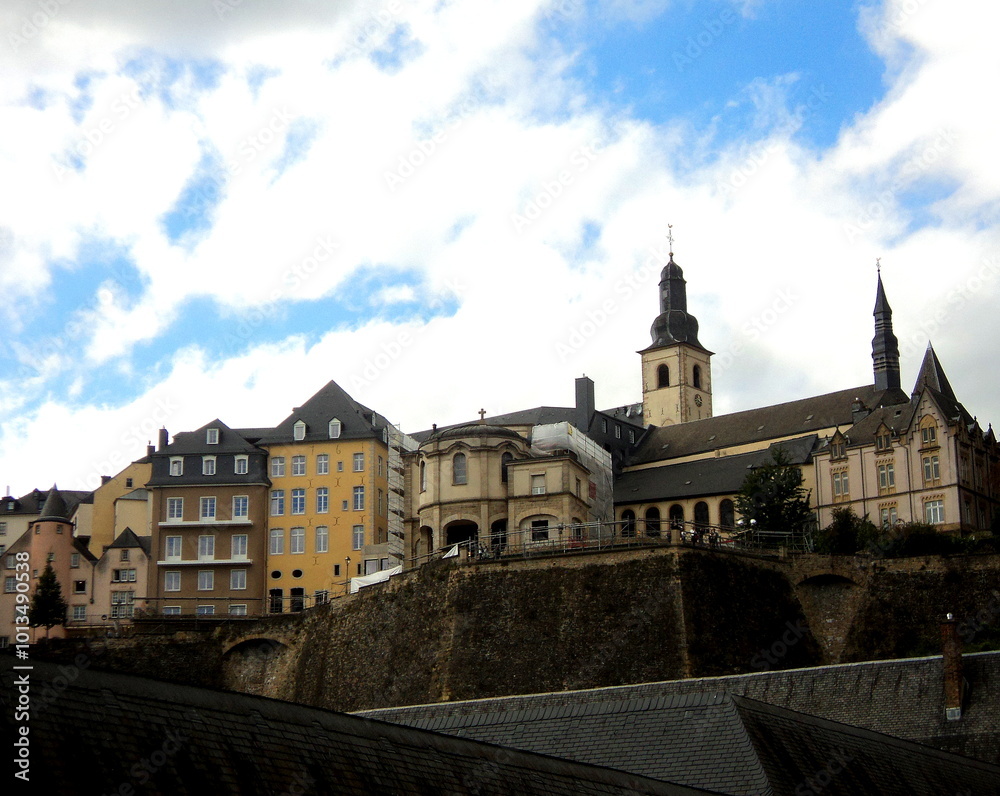 Naklejka premium A picturesque view of Luxembourg's Grund, with historic stone buildings, winding cobblestone streets, and lush greenery lining the river beneath towering cliffs.