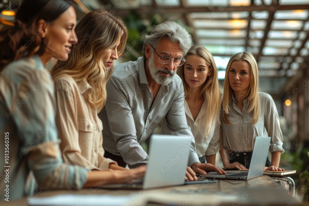 A group of  peopleare standing and smiling at each other as they gather around an open space office table with laptops on it.