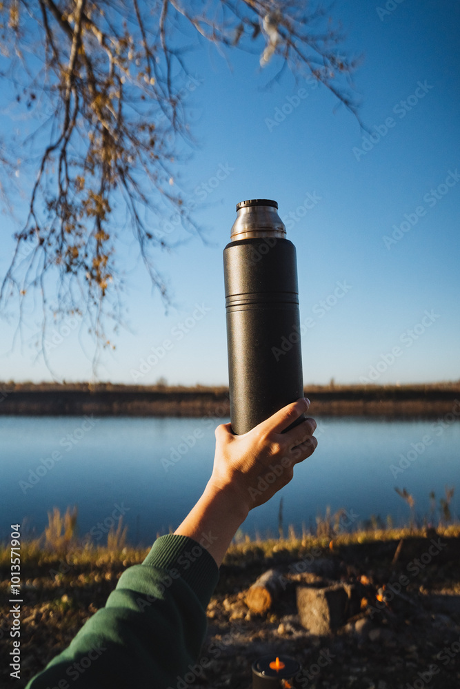 Obraz premium A thoughtful individual is gently holding a thermos while sitting beside a serene lake, fully immersed in the stunning view and the calming atmosphere of nature surrounding them