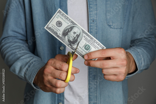 Budgeting. Man cutting dollar banknote with scissors on dark grey background, closeup