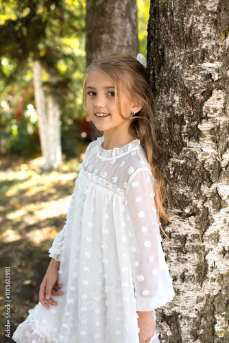 a young girl is standing next to a tree and is wearing a white dress with white polka dots.