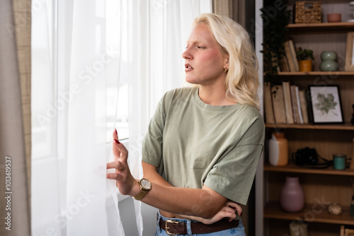 Fotografie Young woman living alone in apartment looking out the window spying on new neighbors moving into house or apartment across the street from hers
