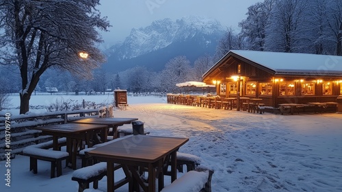 A cozy wooden cabin with snow-covered tables and benches stands in a snowy landscape with mountains in the distance.