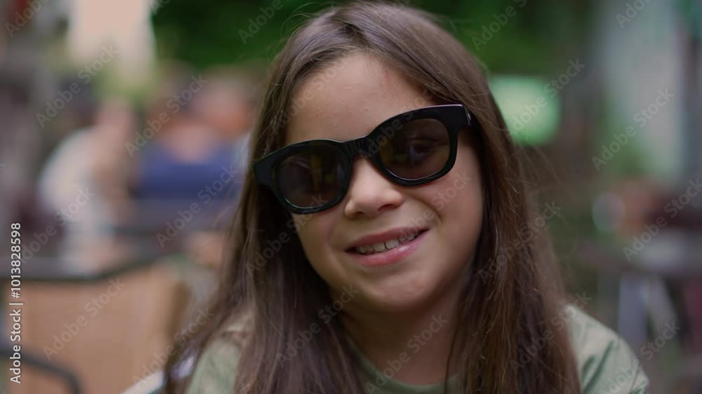 Young girl smiling gently while wearing large sunglasses, sitting comfortably outdoors. conveys a cheerful and carefree moment, embodying childhood joy and innocence