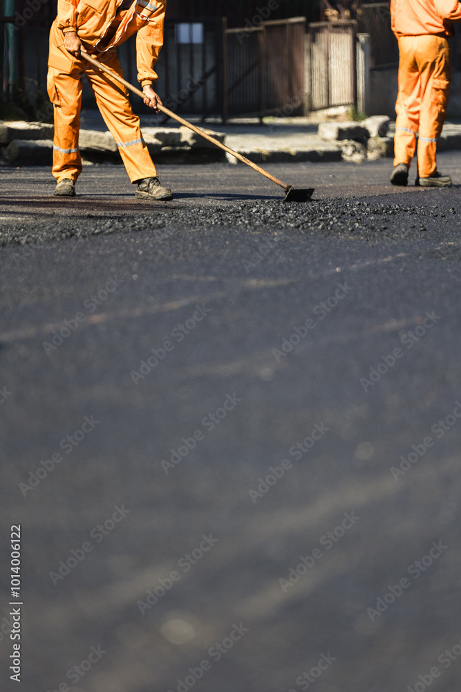 Workers in orange uniforms repair a street by spreading asphalt during the day in an urban setting, showcasing the effort involved in maintaining road infrastructure Workers in orange uniforms repair a street by spreading asphalt during the day in an urban setting, showcasing the effort involved in maintaining road infrastructure