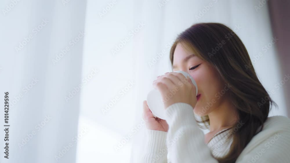Young asian woman standing beside window and holding mug in bedroom at home, She drinking milk after wake up in the morning Young asian woman standing beside window and holding mug in bedroom at home, She drinking milk after wake up in the morning
