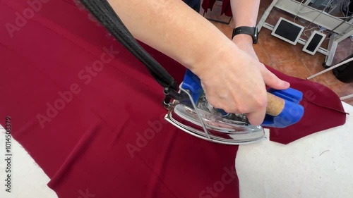 The process of sewing trousers. The hand presses the iron on the red part of the trousers lying flat on the white surface of the ironing table.