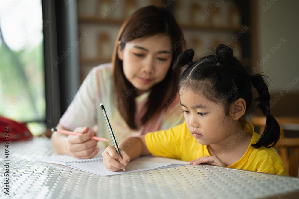 Fototapeta premium A woman is helping a young girl write in a notebook. The girl is wearing a yellow shirt and has her hair in pigtails. The woman is holding a pencil and a pen, and the girl is using a pencil