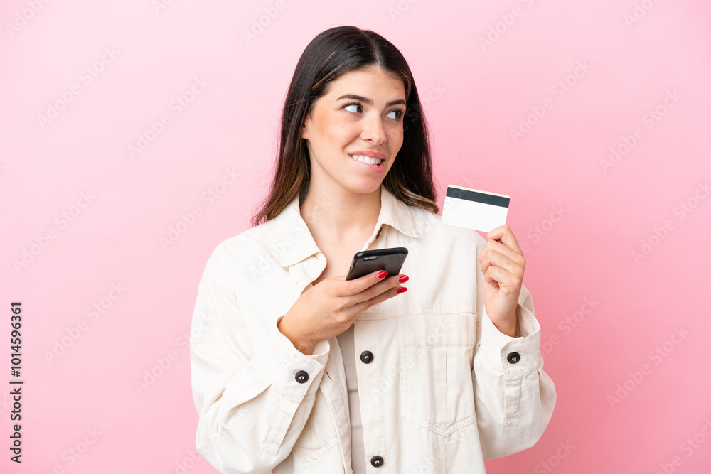 Young Italian woman isolated on pink background buying with the mobile with a credit card while thinking Young Italian woman isolated on pink background buying with the mobile with a credit card while thinking