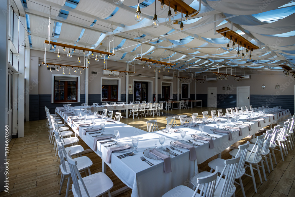 A bright, glass-walled banquet hall with tables covered with white tablecloths. Tableware ready to serve dishes. Photo taken during the day, with sunlight streaming in.