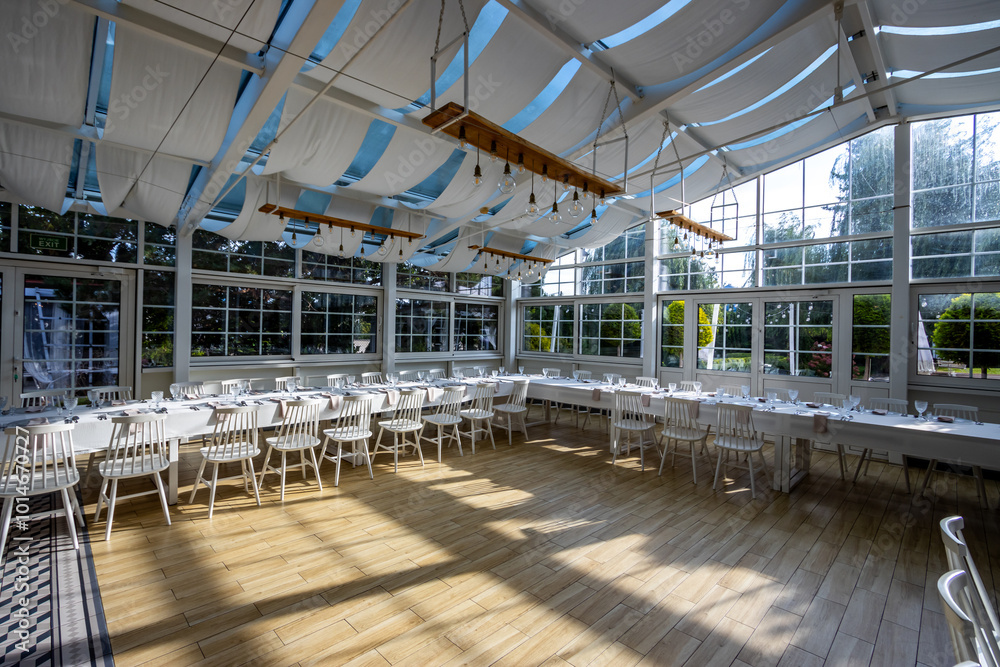 A bright, glass-walled banquet hall with tables covered with white tablecloths. Tableware ready to serve dishes. Photo taken during the day, with sunlight streaming in.