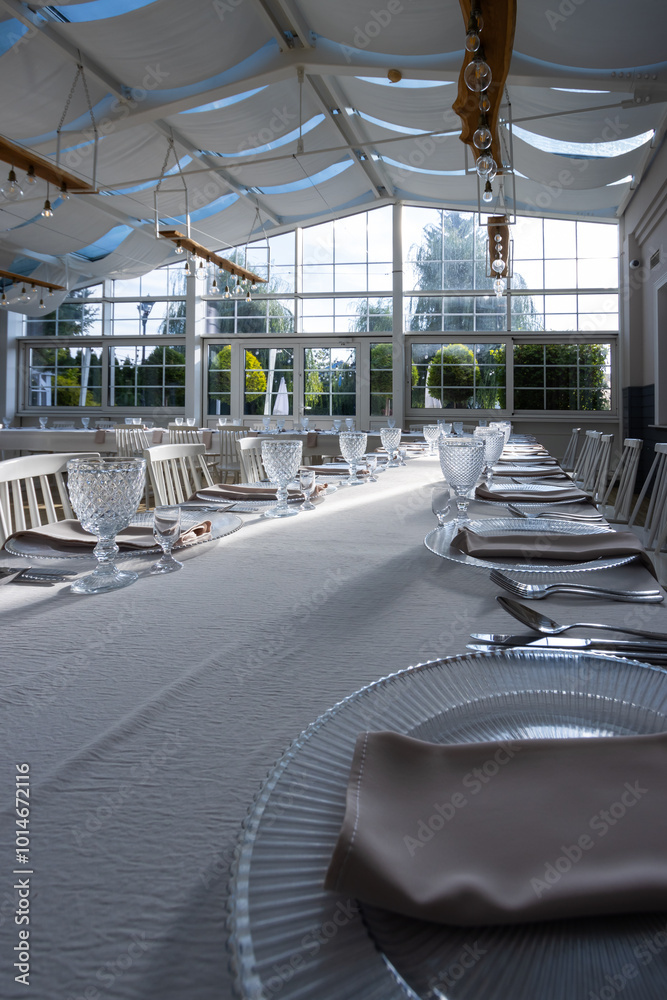 A bright, glass-walled banquet hall with tables covered with white tablecloths. Tableware ready to serve dishes. Photo taken during the day, with sunlight streaming in.