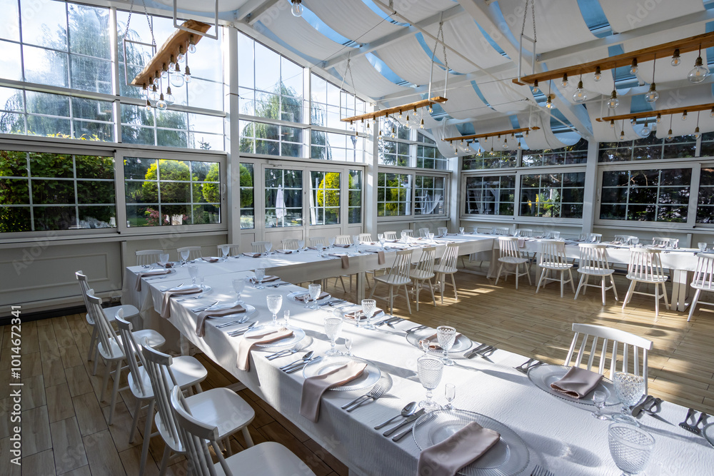 A bright, glass-walled banquet hall with tables covered with white tablecloths. Tableware ready to serve dishes. Photo taken during the day, with sunlight streaming in.