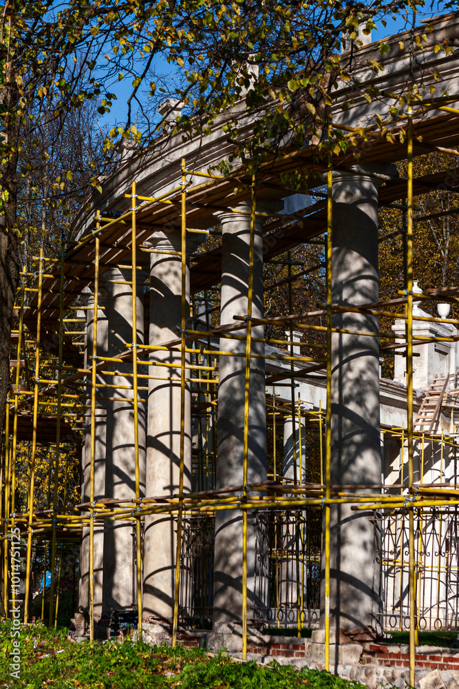 Fototapeta premium The image shows a neoclassical colonnade under restoration, with scaffolding surrounding the white columns. Trees with autumn leaves and a clear blue sky form the natural backdrop.