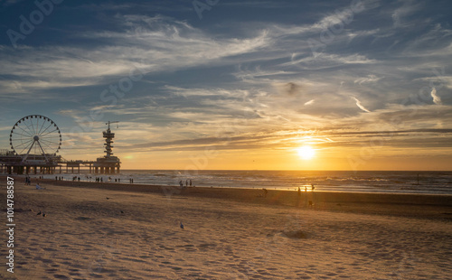 beach at sunset with a Ferris wheel and pier in the distance. The golden sunlight reflects on the ocean waves, while people stroll along the sandy shore, capturing a peaceful, scenic moment