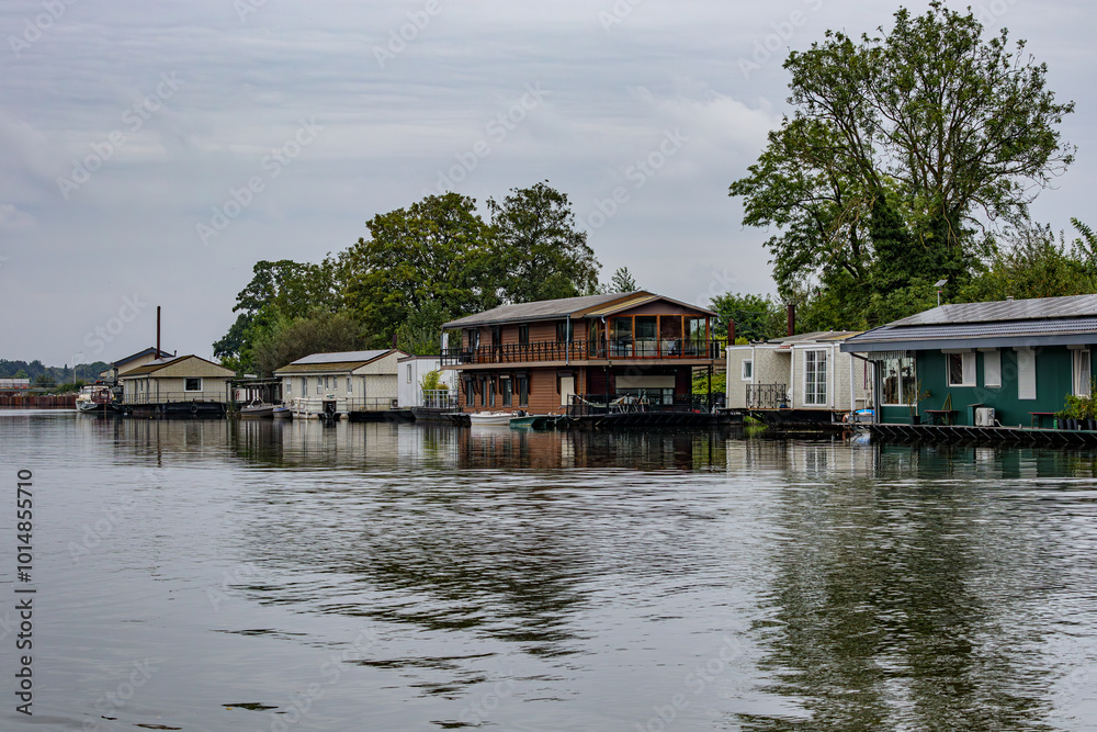 Obraz premium River Meuse with different types of houseboats moored at riverbank, floating cityscape against grey sky, leafy trees in background, cloudy day in Maastricht, South Limburg, Netherlands