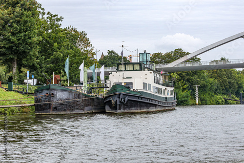 Front view of two boats anchored at small pier on embankment of Meuse river, abundant green leafy trees and bridge in background, cloudy day in Maastricht, South Limburg, Netherlands