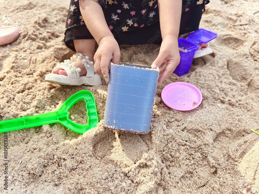 The girl is sitting on the sand and playing sand toys set. A little girl playing beach toys. Kid activity. The kid playing toys in the sandbox The girl is sitting on the sand and playing sand toys set. A little girl playing beach toys. Kid activity. The kid playing toys in the sandbox